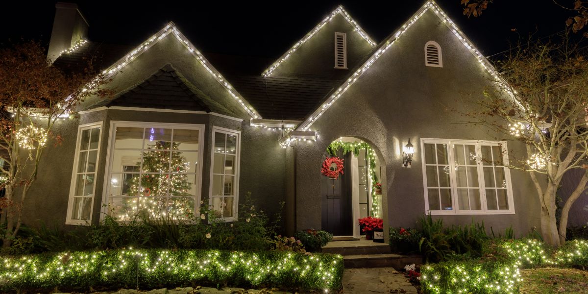 Home exterior at night with Christmas lights outlining the roofline and illuminating front-yard shrubs.